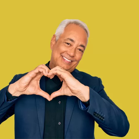 A healthy clinical trial participant in a navy blazer makes a heart with their hands and smiles at the camera against a bright yellow background.