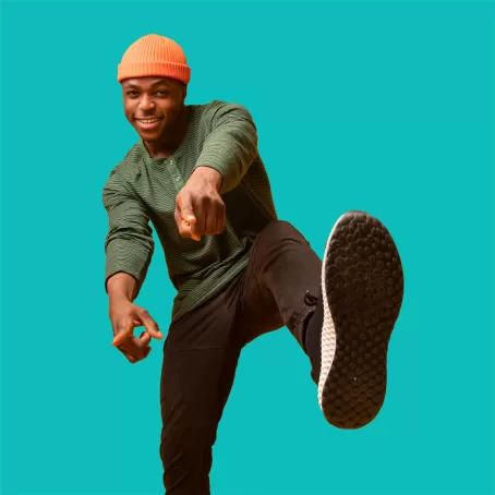 A healthy clinical trial volunteer with an orange beanie stands on one leg and points at the camera against a bright teal background.