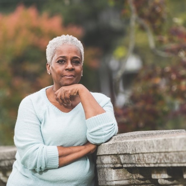 Retrato de una mujer afroamericana mayor, parada en un parque, apoyada en una barandilla de piedra con árboles desenfocados al fondo. Está mirando hacia otro lado, pensando, con la barbilla apoyada en el dorso de la mano - Pfizer Clinical Trials