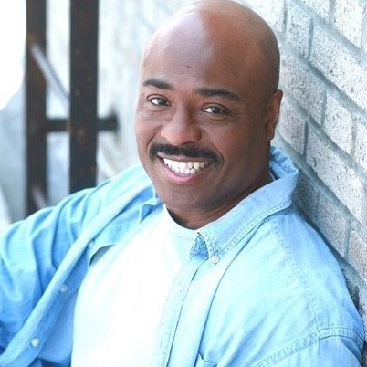 A clinical trial participant sits outside against a white brick wall smiling at the camera