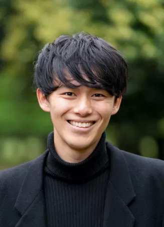 A young Japanese male smiles at the camera with trees in the background