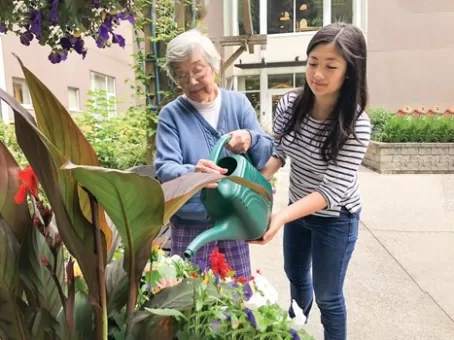 Nieta multiétnica ayudando a la abuela a regar un recipiente con flores en el jardín: estudios clínicos de Pfizer