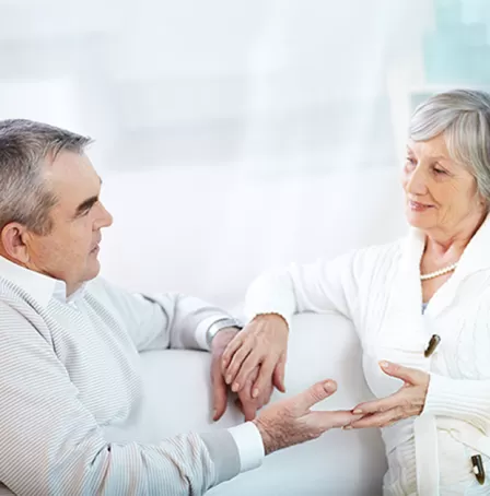 Portrait of a senior male and female couple at home holding hands. Neutral colors used throughout. – Pfizer Clinical Trials