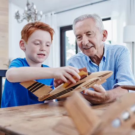 The image in the foreground is of an older man and a young child holding a wooden model plane sat at a table. In the background is a neutrally toned and modern living area.– Pfizer Clinical Trials