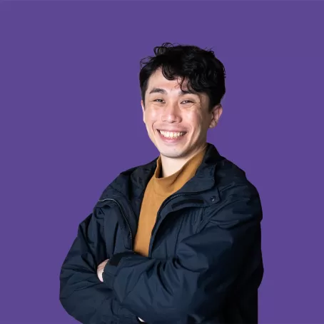 A healthy clinical trial volunteer in a dark blue jacket smiles with their arms across their chest against a bright purple background.