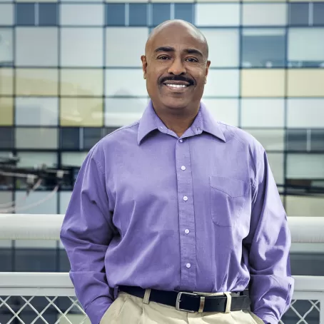 John, a healthy volunteer at the Pfizer CRU in New Haven, stands in a purple shirt smiling on a walkway