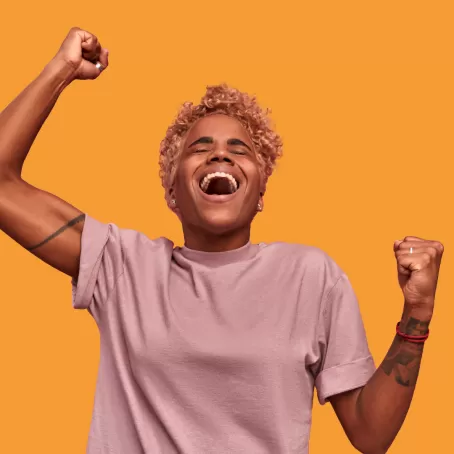healthy clinical trial volunteer in a beige shirt pumps their fists excitedly and laughs against a bright orange background