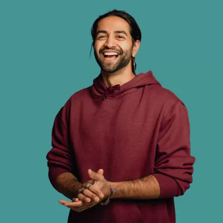 A clinical trial volunteer in a burgundy hoodie clasps their hands and smiles at the camera against a teal background