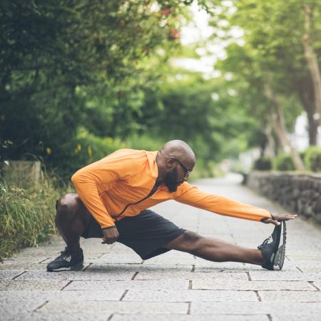 An adult in an orange long-sleeve top stretches their legs on a sidewalk with trees in the background