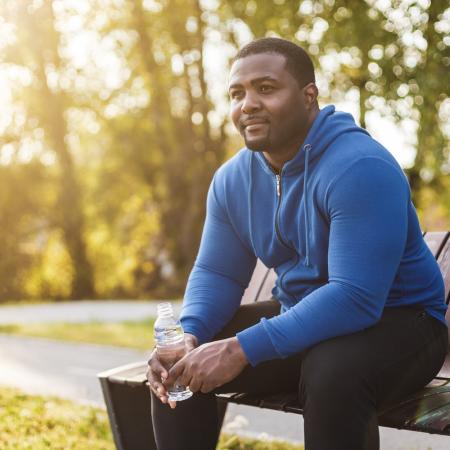 Man in a blue hoodie sitting on a park bench at sunset, holding a water bottle and looking thoughtful – Pfizer Clinical Trials
