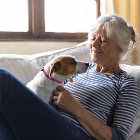 Woman reclines on a couch in a sun lit living room, while playing with her dog who sits on her lap – Pfizer Clinical Trials