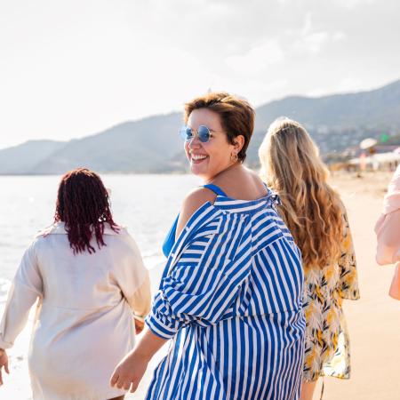 Group of women walking along a sunny beach, with one woman in blue and white stripes smiling back at the camera – Pfizer Clinical Trials