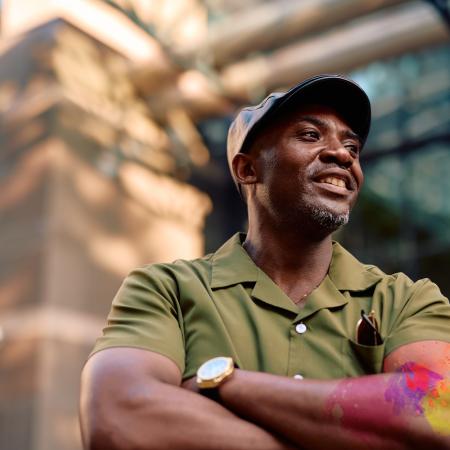 Stylish African American mature man smiling with arms crossed, wearing a green shirt and black cap, enjoying the vibrant city atmosphere while looking thoughtfully away – Pfizer Clinical Trials