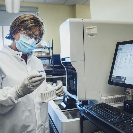 A researcher wearing a lab coat and other PPE checks a sample in front of a computer and other lab equipment during a clinical trial at the PCRU.